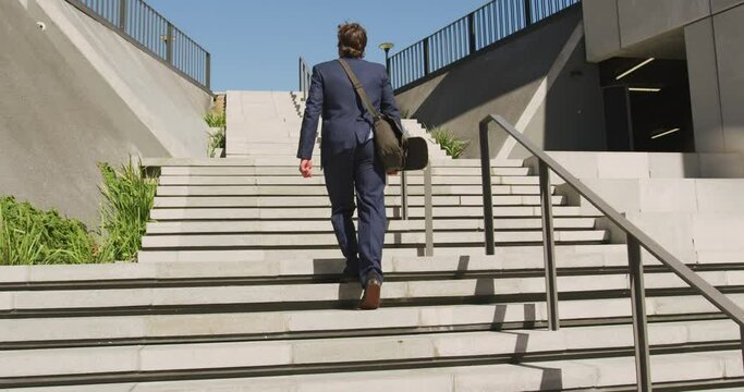 Back view of caucasian businessman walking upstairs, holding skateboard on sunny day
