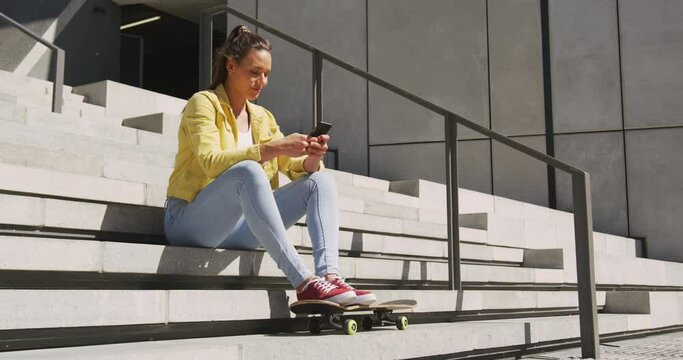 Caucasian woman sitting on stairs with skateboard, using smartphone on sunny day