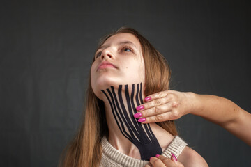 Therapist hands is applying black striped kinesiology tape on the neck of a young caucasian teenager girl on gray background. Aesthetic taping. Salon or home procedures, closeup view 