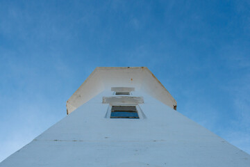The upward view of the watch room or top of a lighthouse that is octagon shaped. There are two small windows in the stark white concrete building with blue sky is in the background. 