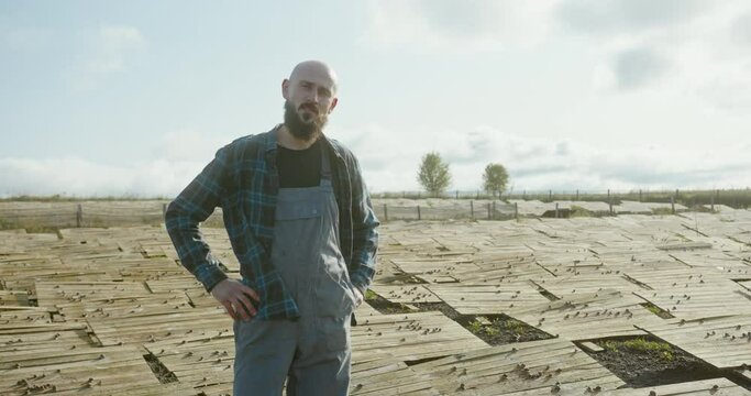 Farmer standing surveying his snail farm looking at the breeding trays outdoors in a field covered in fresh snails looking aside with hands on hips