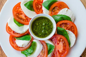 Restaurant menu. Sliced tomatoes with mozzarella cheese, basil leaves and pesto sauce in a white plate against a natural wood background close-up