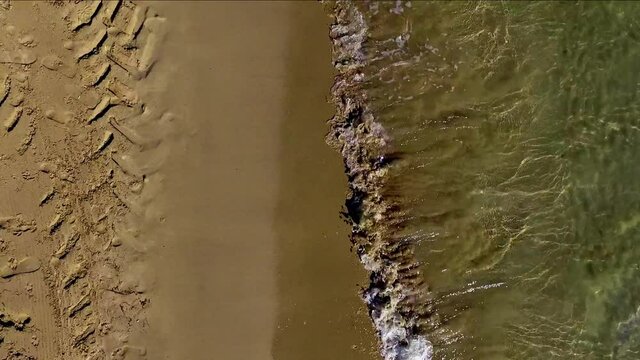Aerial moving view along the sea shore. The waves come and go into the sand. Truck marks are seen on the dry sand.