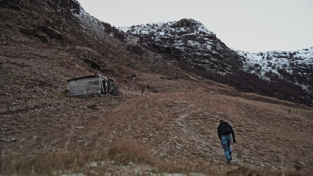 Young man hiking through abandoned meadow with small wooden shed and snow-covered mountains in the background.