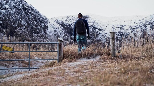 Young Man Hiking Through Meadow And Snow-covered Mountain Terrain, Stepping Over A Fence And Headed Into Mountains, Hoddevik, Norway.