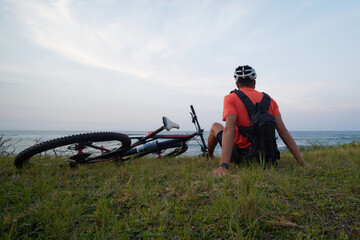The cyclist with backpack, enjoys the view of the ocean, sits on the grass with a bicycle. Back view.