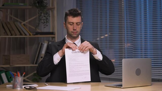 Businessman ripping contract into small apart and throws into air. Man in black business suit sits at table in office. Terminating contract, breaking business agreement, rip deal. Slow motion.