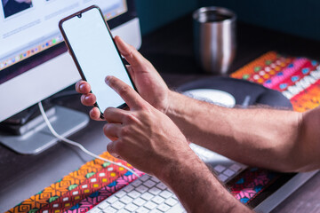Visible hands of a young adult man working at home in front of the computer while holding his smartphone with its blank screen with both hands