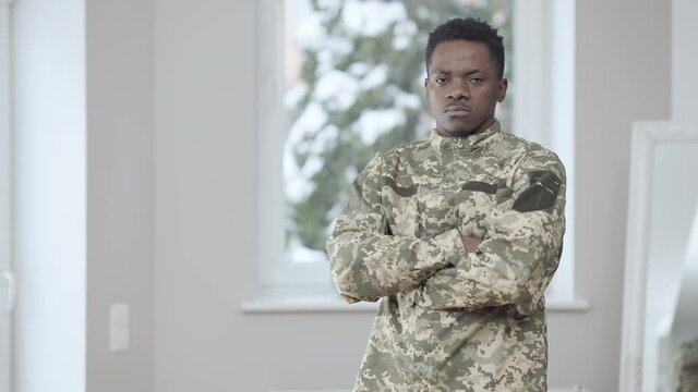 Middle shot of patriotic brave African American man in military uniform crossing hands looking at camera with serious facial expression and smiling. Portrait of proud soldier posing indoors