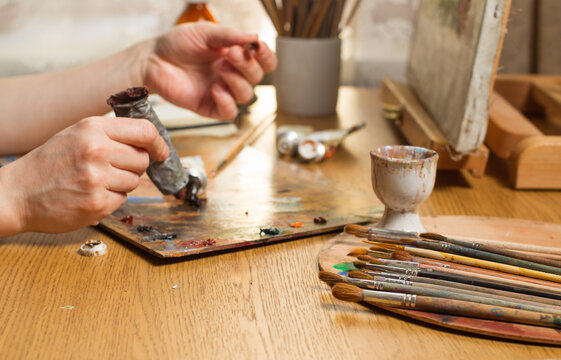 The artist's hands squeeze the paint out of the tube onto the palette. Focus on the bunch of brushes in the foreground