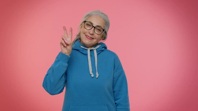 Mature old gray-haired granny grandmother showing victory sign, hoping for success and win, doing peace gesture and smiling with kind optimistic expression. Senior woman isolated on pink background
