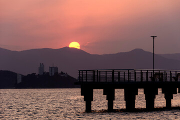 Obraz premium Sunset in the bay of the city of Santos. Silhouette of Fisherman's Pier, Porchat Island and part of the Sun behind the mountains of the Serra do Mar.