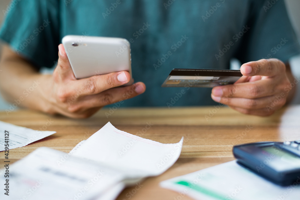 Wall mural Young man wearing wear casual clothes hand holding smart phone and credit card paying bills related to home expenses such as water and electricity bills with calculator bill on wooden office desk.