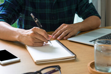 Business man using a laptop to file taxes and write details in a book on wooden office desk at home.
