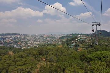 The cable car runs over the coniferous forest. In the distance, in the valley, town houses are visible. There are picturesque cumulus clouds in the blue sky. Dalat. Vietnam