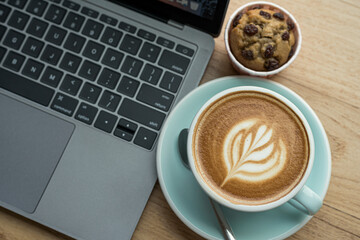 Close-up of Hot coffee latte with latte art milk foam in cup mug and Homemade Banana cup cake with laptop computer on wood desk office desk in coffee shop at the cafe,during business work concept