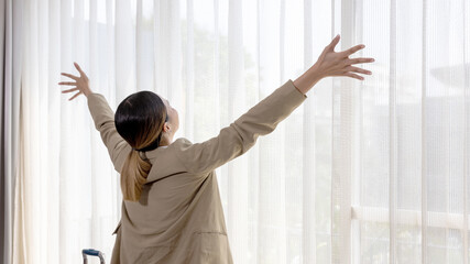 Young Asian girl in smart casual dress takes a breath moment at an apartment room window to inspire relax and stretch. Female visitor or traveler stands in front of white curtain look out for future.