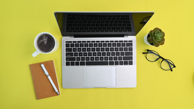 Above view of laptop computer, coffee cup, glasses and notebook on yellow background.