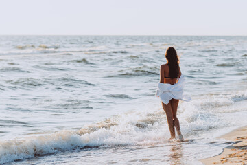 Young beautiful slender woman in a black bikini and white shirt on a tanned body on the beach in the waves, back view. Soft selective focus, art nose.