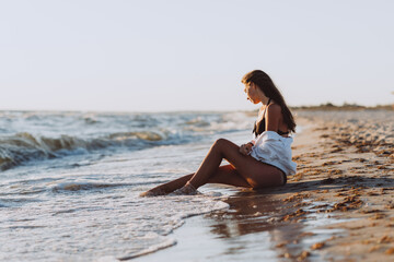 A young beautiful woman in a black bikini and a white shirt on a tanned body sits on the beach on the wet sand. Soft selective focus, art nose.