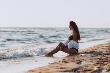 Fototapeta na wymiar A young beautiful woman in a black bikini and a white shirt on a tanned body sits on the beach on the wet sand. Soft selective focus, art nose.