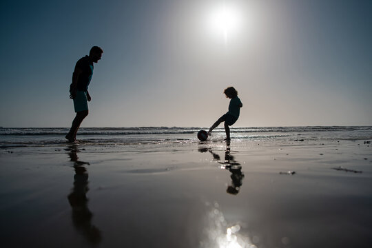 Father and son play soccer or football on the beach on summer family holidays.