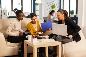 Diverse group of business women sitting on couch in modern corporate startup company office talking about start up financial project and strategy management, using laptop computer and graphics