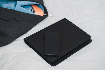 Grey business suit jacket and blue shirt on a wooden hanger and black folder and smart phone laying on a white bed sheet . Getting ready for work and travel concept.