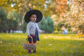 Little girl in witch costume at Halloween in autumn park with basket full of yellow leaves....