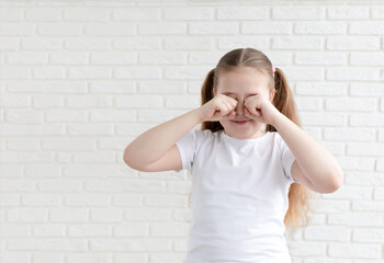 A girl in a white T-shirt covers her eyes with her hands and scratches. The concept of allergy, pollinosis. White brick background. Copy space.
