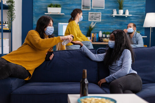 Group Of Multi Ethnic People Using Hand Sanitizer Gel To Prevent Covid19 Spread Spending Time Together In Living Room Sitting On Sofa Wearing Face Mask. Diverse People Socializing Keeping Social