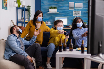 Concentrated multiracial friends playing video games with joystick controller in living room wearing face mask to prevent spreading coronavirus. Diverse people having fun, laughing at new normal party