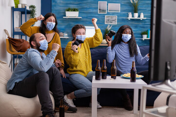 Excited multiracial gamer women sitting on couch playing video games on console during new normal party wearing protective masks. Friends using wireless controllers keeping social distancing