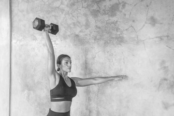 A beautiful female athlete does overhead presses with a dumbbell at a gym