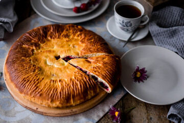 The round pie is cut into pieces. Inside, the filling is made of cherries and sugar. On a wooden table there are dishes, a cup of tea. Berries and flowers are laid out.