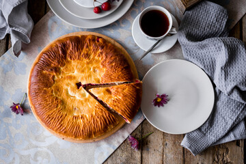 The round pie is cut into pieces. Inside, the filling is made of cherries and sugar. On a wooden table there are dishes, a cup of tea. Berries and flowers are laid out.