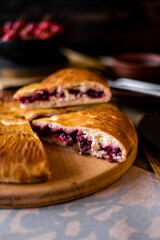 The round pie is cut into pieces. Inside, the filling is made of cherries and sugar. On a wooden table there are dishes, a cup of tea. Berries and flowers are laid out.