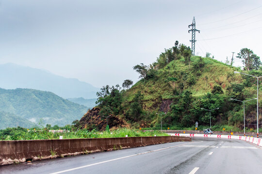 The image of the road that cuts across the mountain covered with rocks, grass, and trees. And there are mountains in the background that line up and fade in the distance.