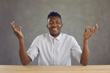 Business pause, take break, mental balance. Happy calm young african american man meditating with eyes closed smiling sitting at desk. Studio headshot face portrait isolated on grey studio background