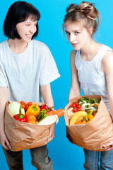 Portrait of Two Funny Caucasian Girls Posing With Eco Paper Bags Filled With Grocery And Vegetables Over Clear Blue Background.