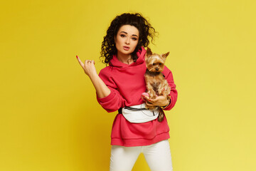 Model girl with afro hair wearing sportswear posing with a small cute dog on her hands over yellow background, isolated