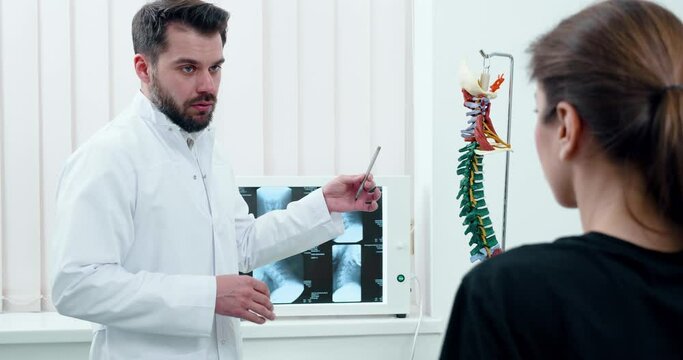 Attractive Man Therapist showing to Patient Girl 3d Model of spine in cabinet. Sucessful Doctor talking with Patient about future Operation while standing in Modern Clinic. X-ray photo. Treatment.