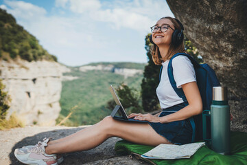 positive young millennial girl with headphones sitting outdoors in mountains working from distance using laptop, beautiful tourist girl having an online video meeting via computer