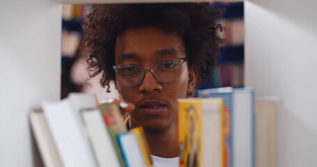 Afro-american male student picking book from bookshelf in library.