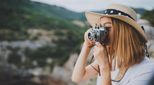 young blonde girl in summer hat takes photo on retro camera on background panorama horizin mountain landscape, hipster tourist enjoys hobby of photographer  leisure in summer nature empty space