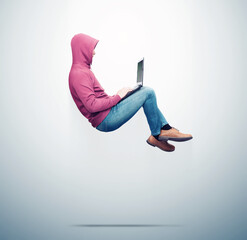 A man in a red sweatshirt, jeans levitates in the air working on a laptop, on blue background. Side...