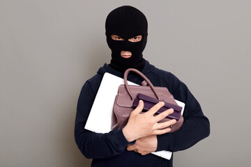 Serious male thief holding bunch of stolen things, laptop, wallet and woman's handbag, robber wearing mask and black turtleneck, burglar isolated over gray background.