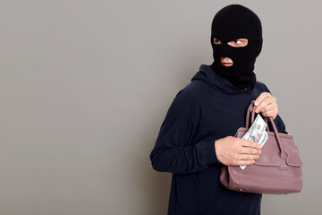 Concentrated thief dresses in balaclava and black turtleneck looks back and holds money and stolen woman's bag in his hands, escapes after robbery and afraid of persecution, isolated on gray wall.