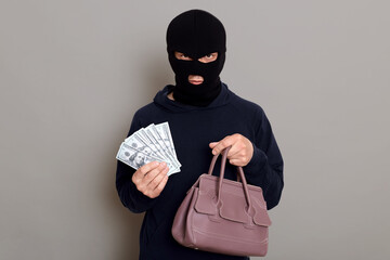 Angry serious thief dressed in balaclava and black turtleneck looks at camera and holds money and...