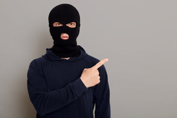 Male thief dressed in black turtleneck and robbery mask points his index finger to side, copy space...
